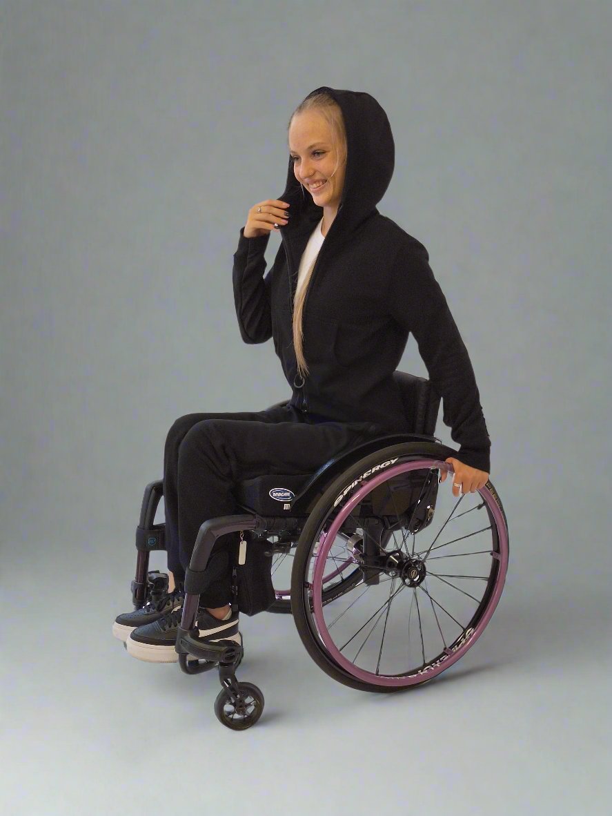 Young woman smiling while seating in a wheelchair. She is wearing black adaptive tracksuit by Vilber's